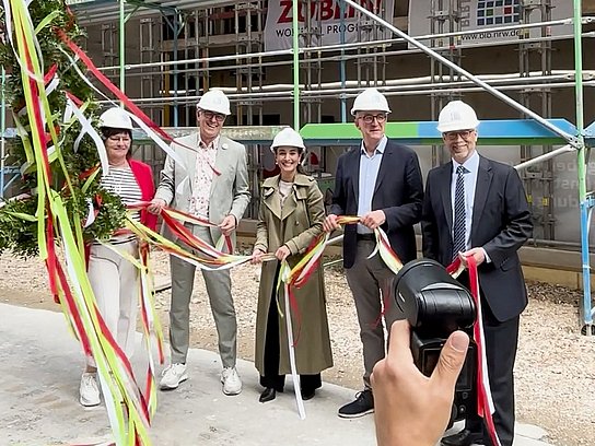 5 Personen stehen vor einer Baustelle, ein Stück des Richtkranz ist zu sehen