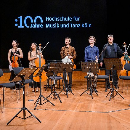 Eine Gruppe von acht Musikerinnen und Musikern steht auf einer Bühne und nimmt den Applaus des Publikums entgegen. Sie halten verschiedene Streichinstrumente wie Violinen, Bratschen und Celli in den Händen.