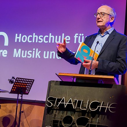 Ein Mann mit Brille spricht an einem Rednerpult mit „STAATLICHE HOCHSCHULE“-Aufschrift. Er hält ein Buch mit der Zahl „10“ und gestikuliert. Im Hintergrund zeigt eine Leinwand „100 Jahre Hochschule für Musik …“. Links steht ein Schlagzeug, die Bühne ist warm beleuchtet.