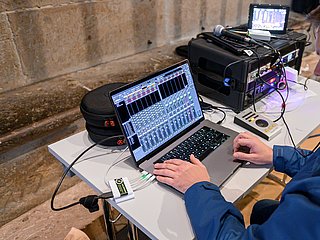  dem Bild sieht man eine Audioproduktionsstation. Eine Person arbeitet an einem Laptop, auf dem eine digitale Audio-Workstation (DAW) mit einem virtuellen Mischpult geöffnet ist. Mehrere Tonspuren und Regler sind auf dem Bildschirm zu sehen. Neben dem Laptop liegen runde Schutzhüllen, und mehrere Kabel sind mit dem Gerät verbunden.