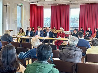 Das Foto zeigt eine Podiumsdiskussion in einem hellen Veranstaltungsraum. Fünf Personen sitzen an einem langen Tisch, der quer im Raum aufgestellt ist. Sie tragen formelle Kleidung und wirken konzentriert. Im Hintergrund sitzt ein kleines Orchester mit Musikerinnen und Musikern, die Streichinstrumente spielen. Der Raum ist mit großen Fenstern ausgestattet, durch die Tageslicht einfällt, und roten Vorhängen. Im Vordergrund sieht man das Publikum von hinten, bestehend aus mehreren Personen, die aufmerksam zuhören. Die Atmosphäre wirkt professionell und zugleich freundlich.