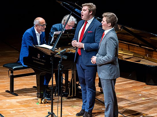 Zwei Sänger stehen vor einem Flügel an dem zwei Pianisten sitzen und gleichzeitig spielen