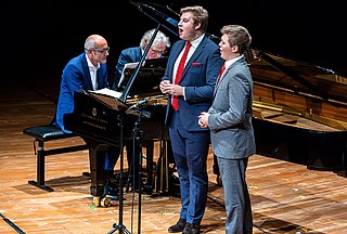 Zwei Sänger stehen vor einem Flügel an dem zwei Pianisten sitzen und gleichzeitig spielen