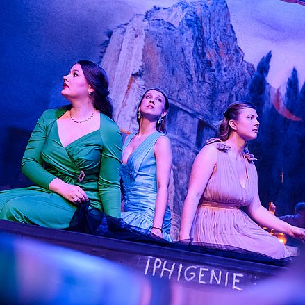 Ariadne auf Naxos - Richard Strauss Drei Darstellerinnen in eleganten Kleidern sitzen auf einer Bühne mit der Aufschrift „IPHIGENIE“