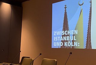 Tisch mit drei Stühlen, Mikrofone auf dem Tisch und an der Wand eine Präsentation "Zwishcne Istanbul und Köln"