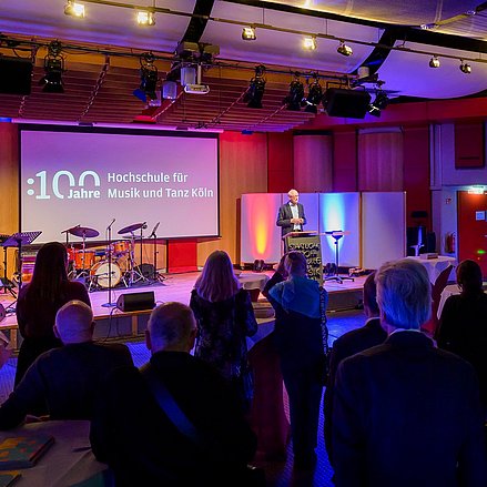 Ein älterer Mann spricht auf einer Bühne an einem Rednerpult. Hinter ihm zeigt eine Leinwand den Schriftzug „100 Jahre Hochschule für Musik und Tanz Köln“. Links auf der Bühne stehen ein Schlagzeug und weitere Instrumente. Im Vordergrund hört ein Publikum aus festlich gekleideten Gästen aufmerksam zu, einige stehen an Stehtischen. Der Raum ist in warmes und farbiges Licht getaucht.